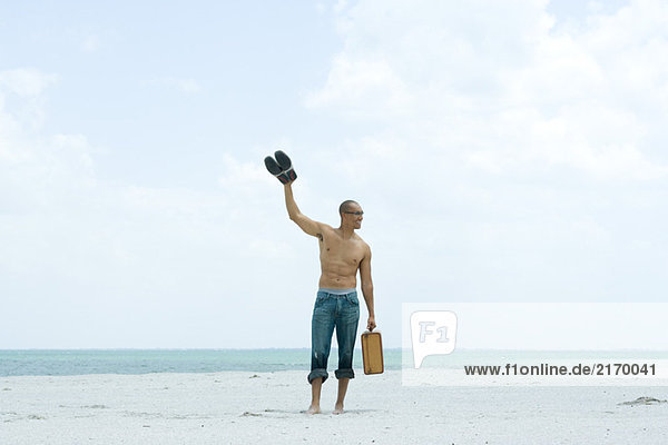 Man walking on beach carrying suitcase and waving  looking away
