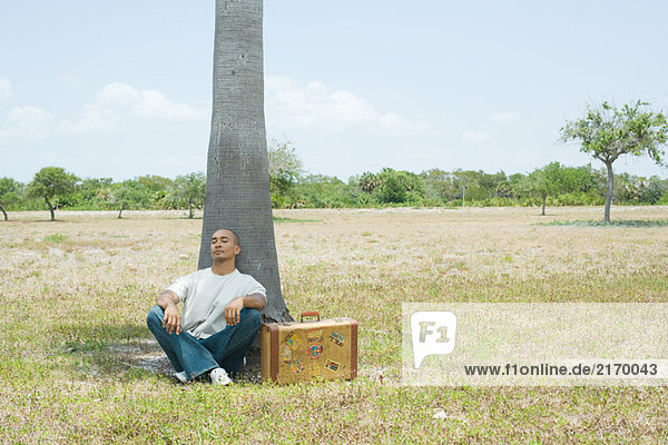 Man with suitcase sitting on the ground  leaning against tree with eyes closed