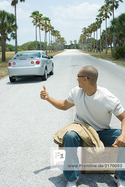 Male hitchhiker sitting on suitcase on road  looking over shoulder at passing car