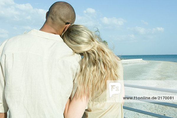 Couple standing arm in arm looking at view  woman's head on man's shoulder  rear view