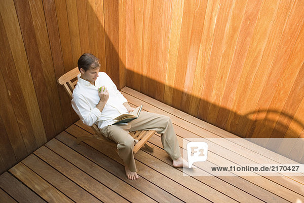 Man sitting in lounge chair reading book  apple in hand  high angle view
