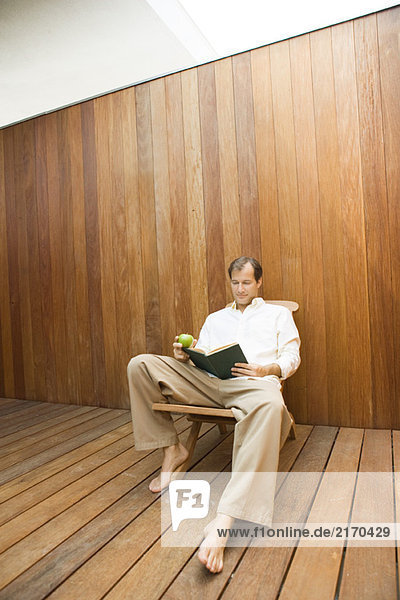 Man sitting in lounge chair reading book  apple in hand