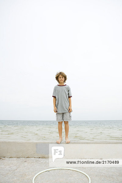 Junge steht auf einer niedrigen Wand am Strand und schaut in die Kamera.