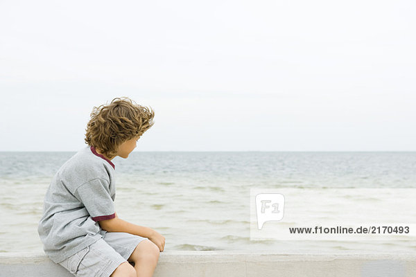 Junge sitzt auf einer niedrigen Wand am Strand und schaut auf die Aussicht.