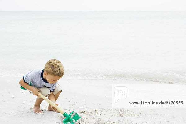 Young boy crouching at the beach  digging in sand with shovel