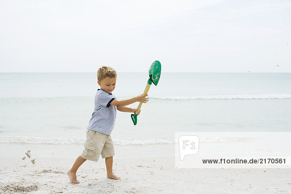 Young boy standing on beach  holding up shovel  looking down