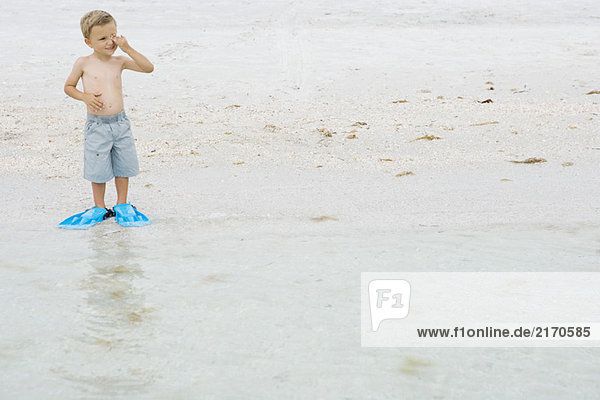 Kleiner Junge mit Flossen am Strand  steht am Wasserrand  reibt sich die Augen