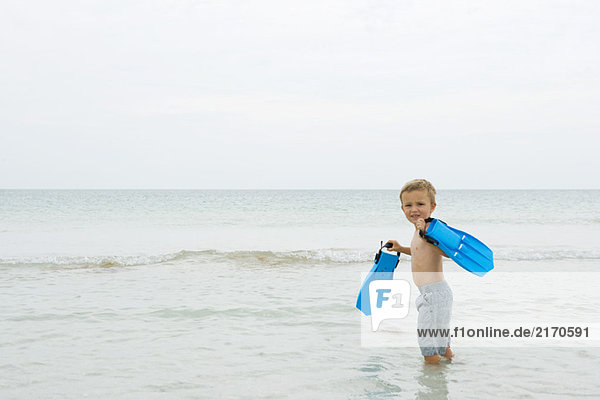 Young boy standing knee deep in water  carrying flippers  smiling over shoulder at camera