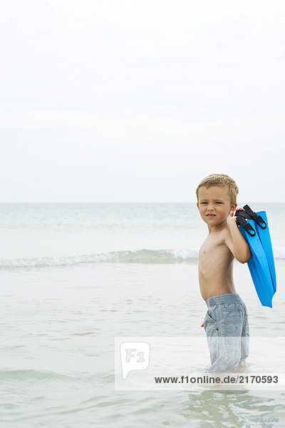 Kleiner Junge steht knietief im Wasser  trägt Flossen  schaut über die Schulter zur Kamera.