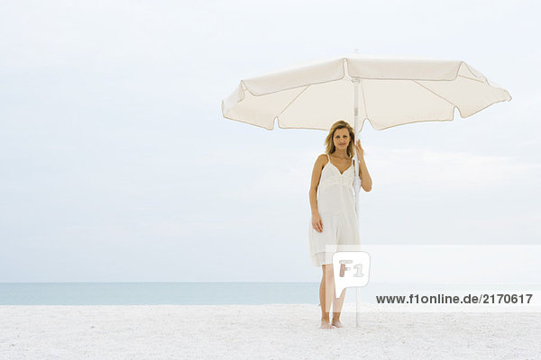 Frau im Sonnenkleid steht unter dem Sonnenschirm am Strand und schaut in die Kamera.