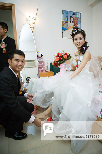 Groom putting shoe on bride's foot in bedroom  both smiling at camera  full length