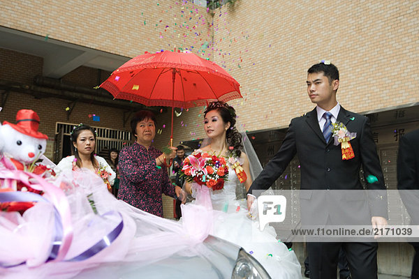 Chinese wedding  bride and groom leaving under confetti  bride covered by red parasol  decorated car in foreground