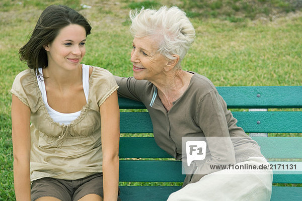 Großmutter und jugendliche Enkelin sitzen zusammen auf der Bank  beide lächelnd.