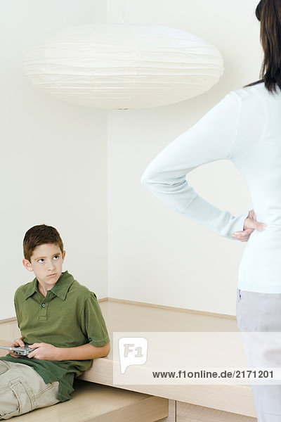 Junge mit einem Videospiel  das die Mutter anstarrt  die ihn mit den Händen auf den Hüften beobachtet.