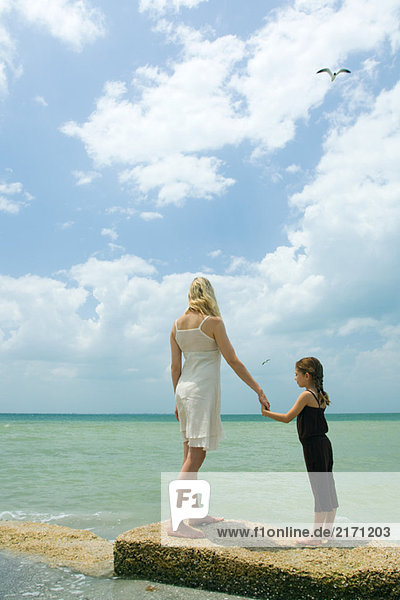 Mutter und Tochter stehen auf dem Wellenbrecher  halten sich an den Händen und blicken auf den Ozean.