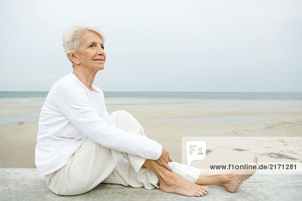 Seniorin auf niedriger Wand sitzend,  Strand im Hintergrund,  volle Länge