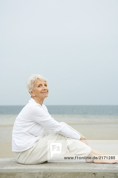 Barfuß Seniorin auf niedriger Wand sitzend  lächelnd  Strand im Hintergrund  volle Länge