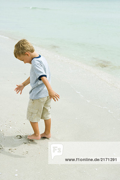 Kleiner Junge  der am Strand steht und Zehen in Sand gräbt.