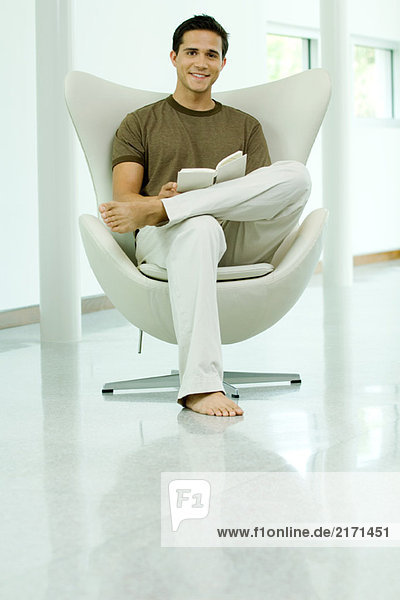 Young man sitting in chair holding book  smiling at camera  full length