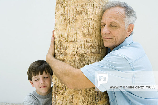 Grandfather and grandson hugging tree trunk  eyes closed