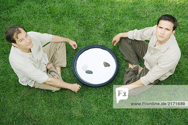 Zwei Männer auf dem Boden sitzend  mit Steingarten dazwischen  mit Blick auf die Kamera  hohe Blickwinkel