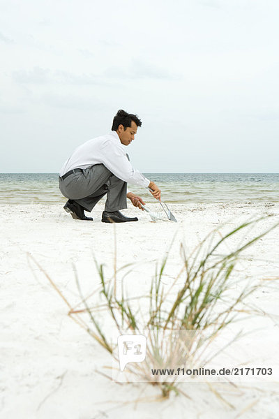 Mann hockt am Strand  fegt Sand in die Kehrschaufel  Seitenansicht