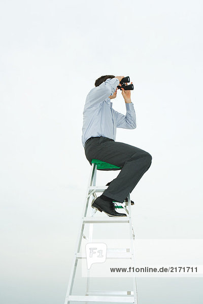 Mann auf der Leiter sitzend durchs Fernglas schauend  Seitenansicht