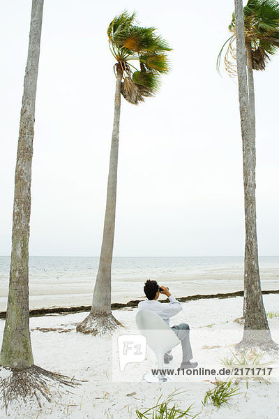 Mann im Stuhl am Strand sitzend  durchs Fernglas schauend  Rückansicht
