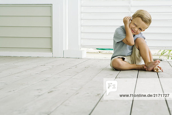 Boy sitting on the ground listening to seashell  squinting at camera  smiling