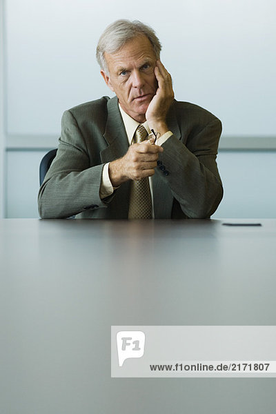 Businessman sitting with hand under chin  holding glasses in hand  looking at camera