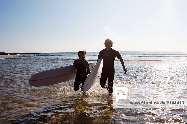 Ein Paar  das lächelnd durchs Wasser läuft und Surfbretter trägt.