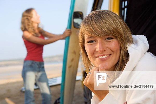 Frau am Strand lächelnd mit einer anderen Frau im Hintergrund die das Surfbrett hält.