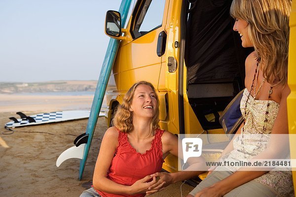 Zwei Frauen sitzen bei einem Van und lächeln mit Surfbrettern im Hintergrund.