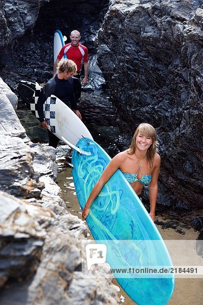 Drei Leute tragen Surfbretter an großen Felsen lächelnd.