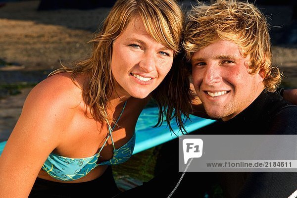 Paar sitzend zusammen lächelnd mit Surfbrett im Hintergrund.