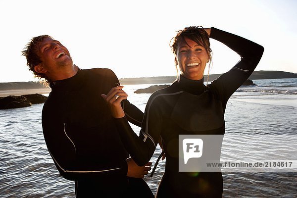 Paar in Neoprenanzügen die am Wasser lachen.