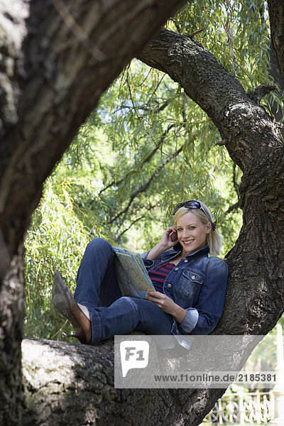 Frau sitzt auf einem Baum auf dem Handy und lächelt.