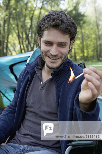 Man sitting at campsite holding a lit match smiling.