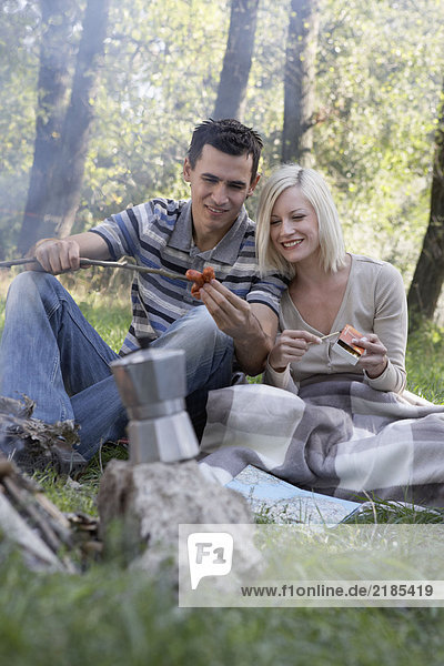 Paar auf dem Campingplatz kocht Hot Dogs und lächelt.