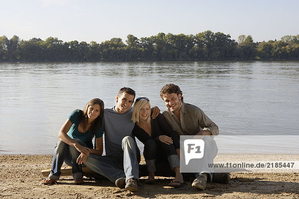 Vier Freunde sitzen auf einem Baumstamm am See und lächeln.