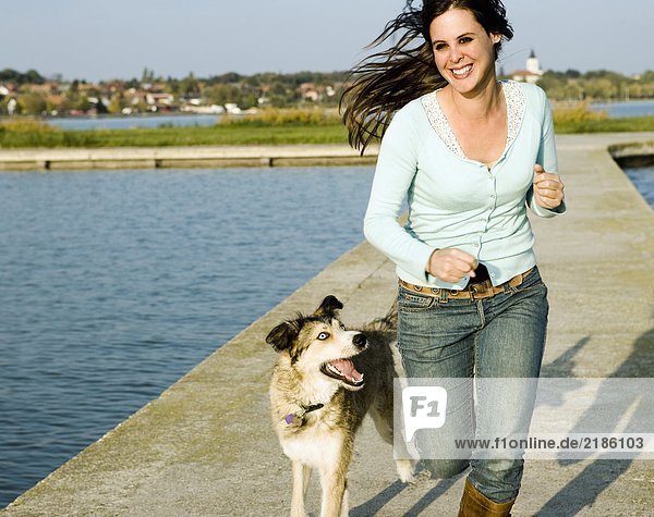 Frau läuft mit Hund an einem See entlang.