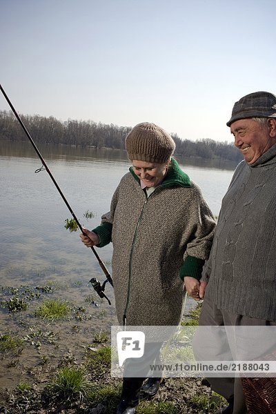 Senior couple walking by river  woman carrying fishing rod  smiling