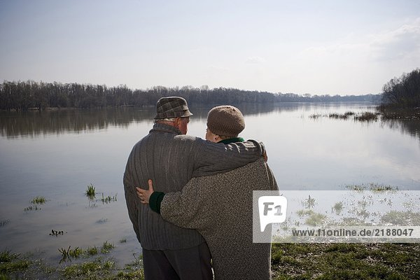 Seniorenpaar steht am Fluss  Arme umeinander  Rückansicht