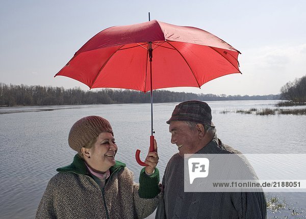 Senior couple standing by river sharing umbrella  smiling
