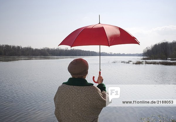 Senior woman standing by river holding red umbrella  rear view