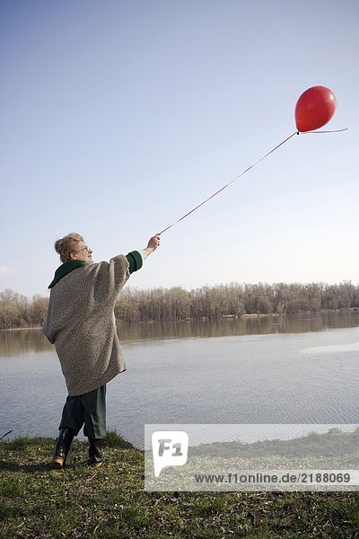 Senior woman standing by river holding red balloon  rear view