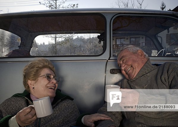 Senior couple sitting by car in countryside  holding cups  smiling