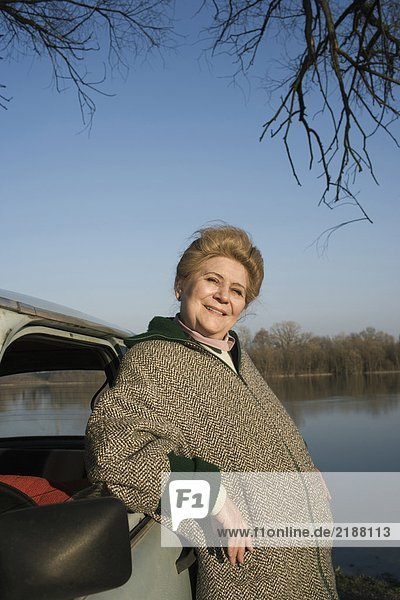 Senior woman leaning on car by riverside  smiling  portrait