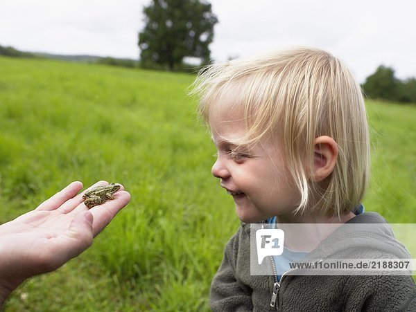 Ein junges Mädchen  das lächelnd auf einen kleinen Frosch in der Hand eines Mannes schaut.
