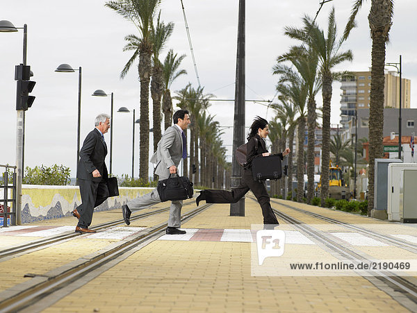Businesswoman and two businessmen running acrossdouble tram lines with suitcases at zebra crossing. Alicante  Spain.